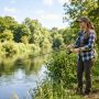A beginner angler holding a spinning rod and reel on a peaceful riverbank on a sunny morning
